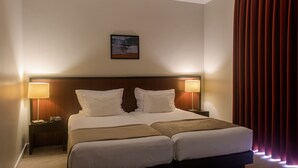 Ein Hotelzimmer mit zwei Betten, einem Kopfteil, Nachttischlampen, einem Nachttisch und einem gerahmten Bild an der Wand.