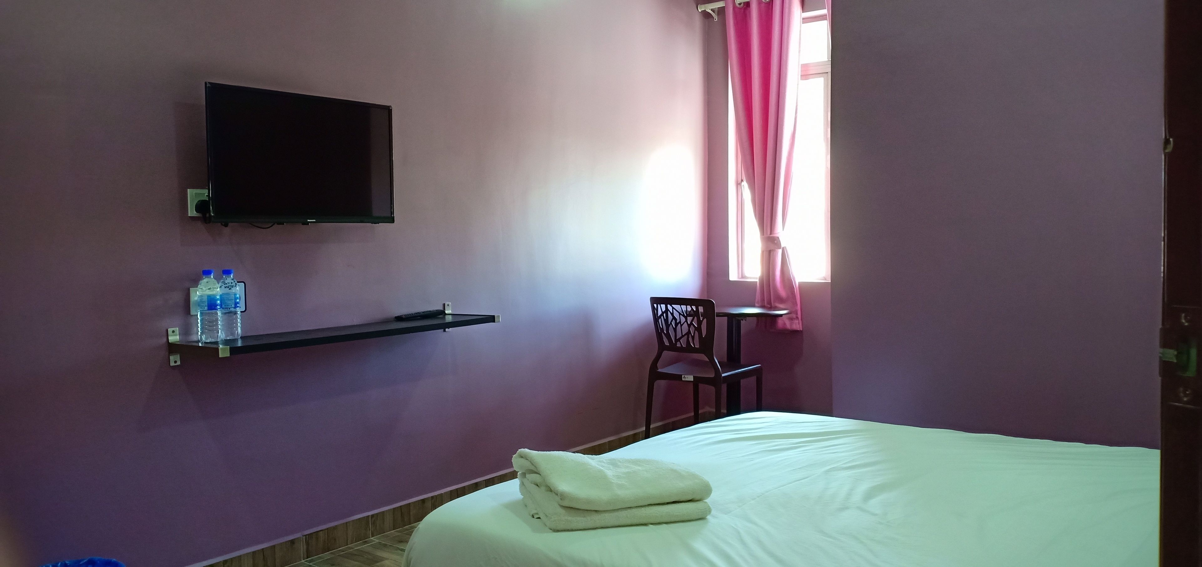Una habitación de hotel con una cama, un televisor montado, una estantería con botellas de agua, una silla y una mesa.