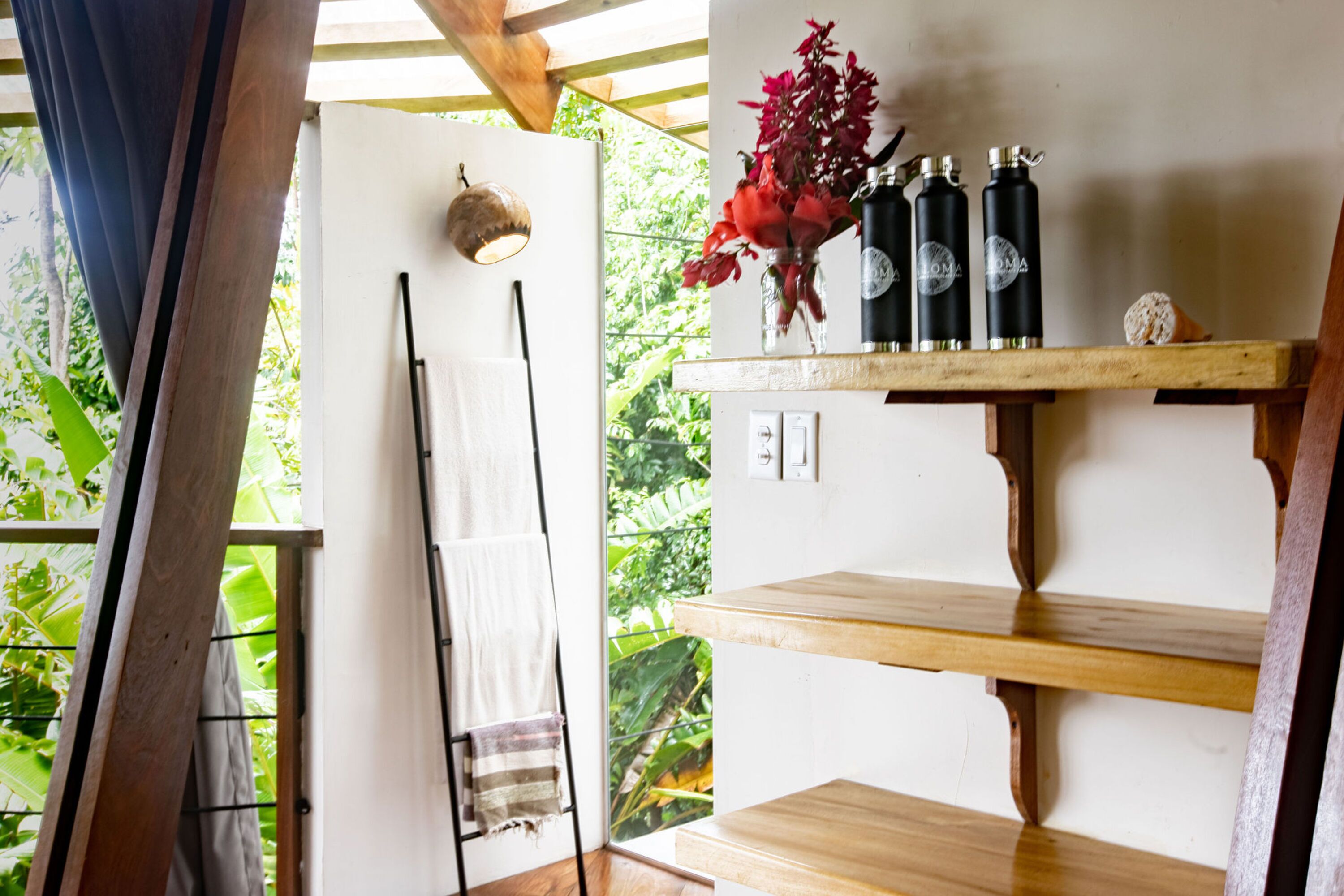 Una habitación con un estante de madera con botellas y un jarrón de flores, una toalla colgada en un perchero y vistas al verdor exterior.
