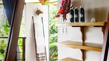 Una habitación con un estante de madera con botellas y un jarrón de flores, una toalla colgada en un perchero y vistas al verdor exterior.