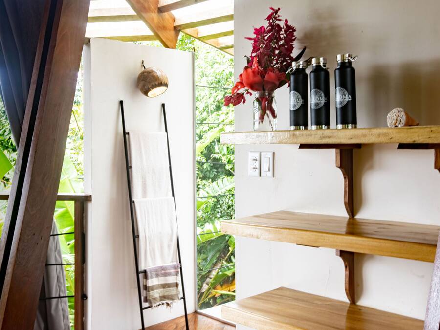 Una habitación con un estante de madera con botellas y un jarrón de flores, una toalla colgada en un perchero y vistas al verdor exterior.