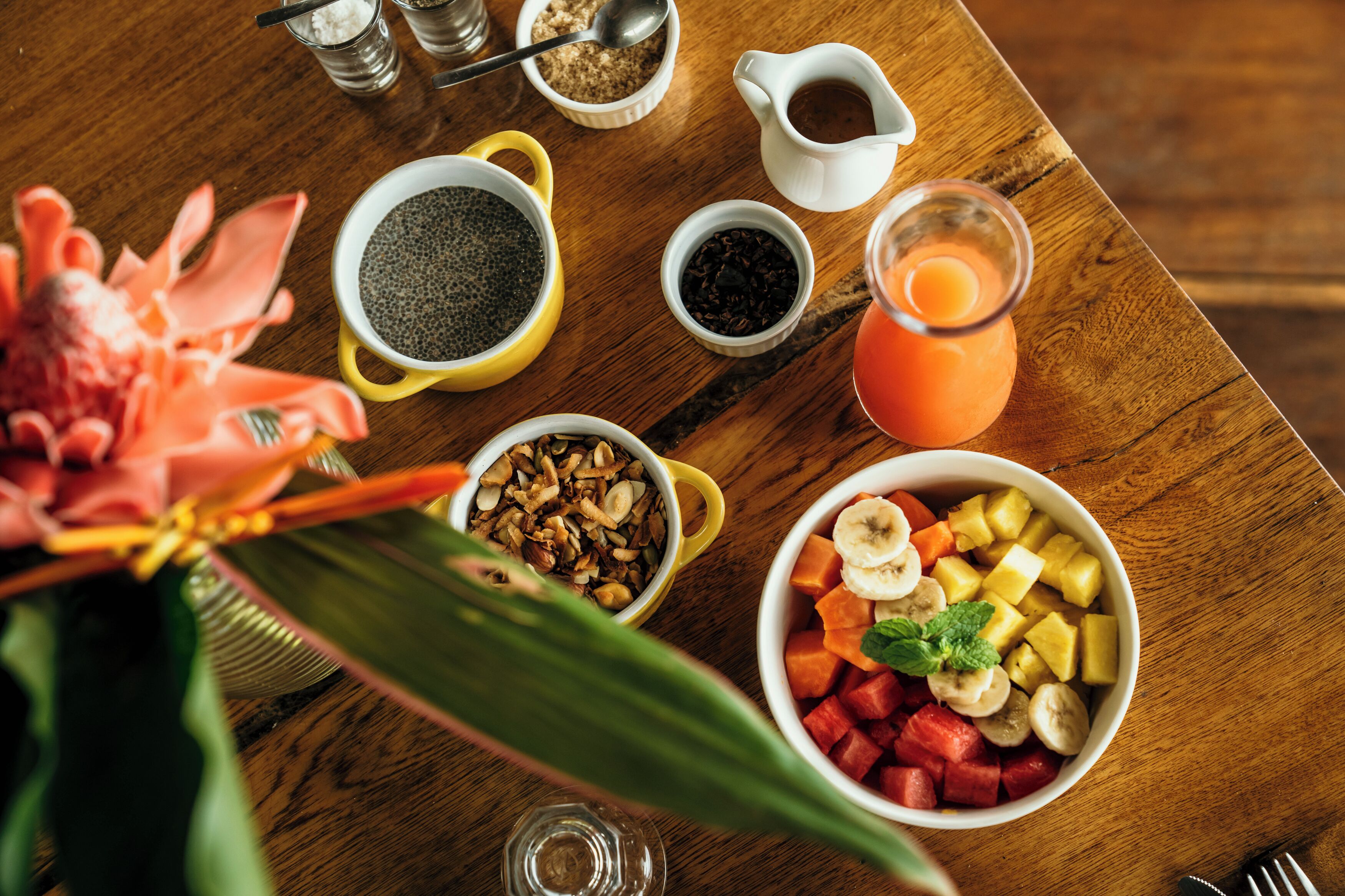 Una mesa de madera con diversos alimentos para el desayuno, incluyendo un tazón de fruta, un tazón de granola, una taza de café y un vaso de jugo de naranja.