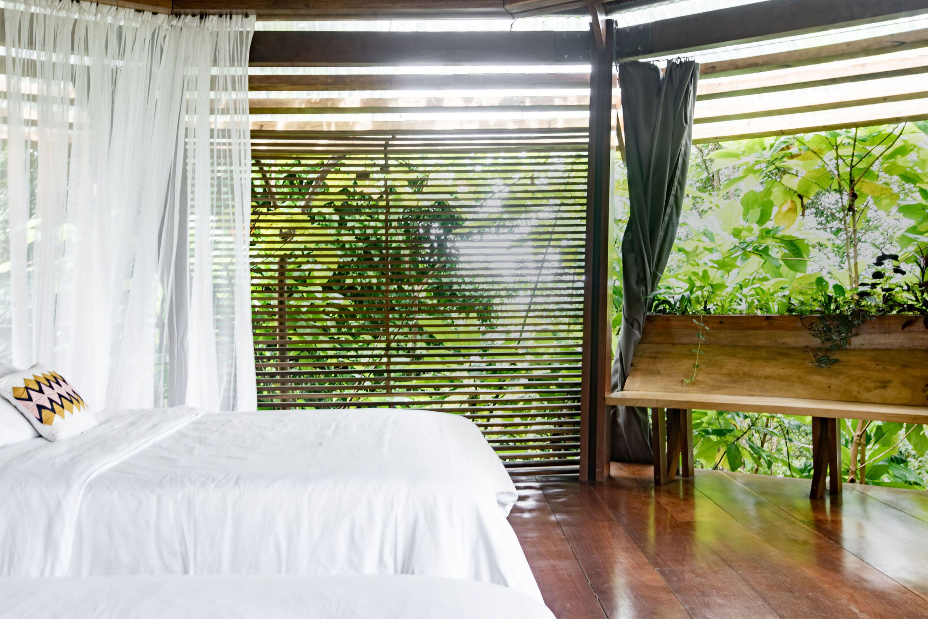A bedroom with a bed, white curtains, a wooden bench, and a view of greenery through bamboo blinds.