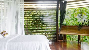 A bedroom with a bed, white curtains, a wooden bench, and a view of greenery through bamboo blinds.