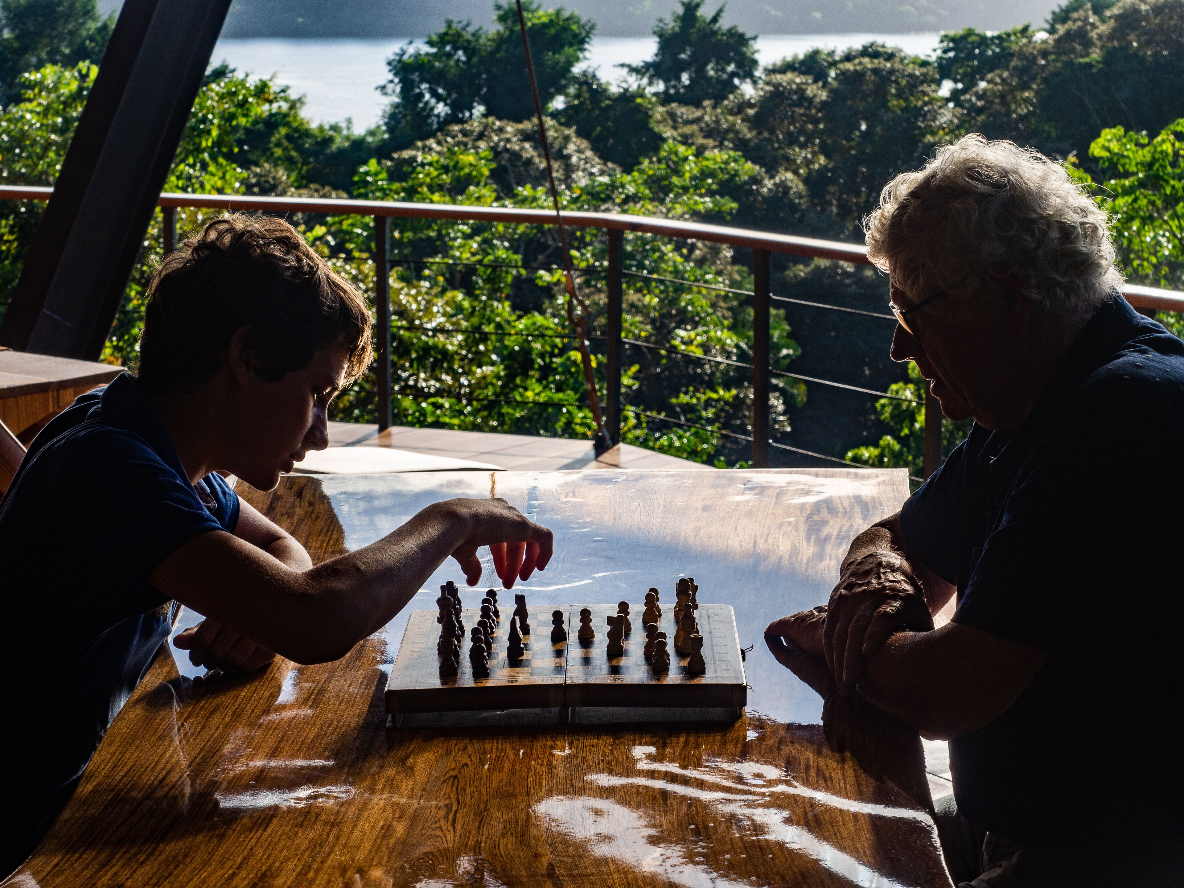 Deux personnes jouent aux échecs sur une table en bois, avec en arrière-plan un paysage pittoresque composé d’arbres et d’eau.