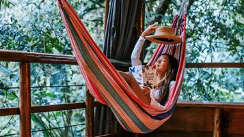 Un fauteuil suspendu en forme de hamac, avec une personne allongée à l’intérieur, une terrasse en bois et une vue sur les arbres.