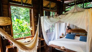 Una habitación de madera con una cama bajo un dosel, una hamaca, una guitarra y una ventana con vistas a la vegetación.