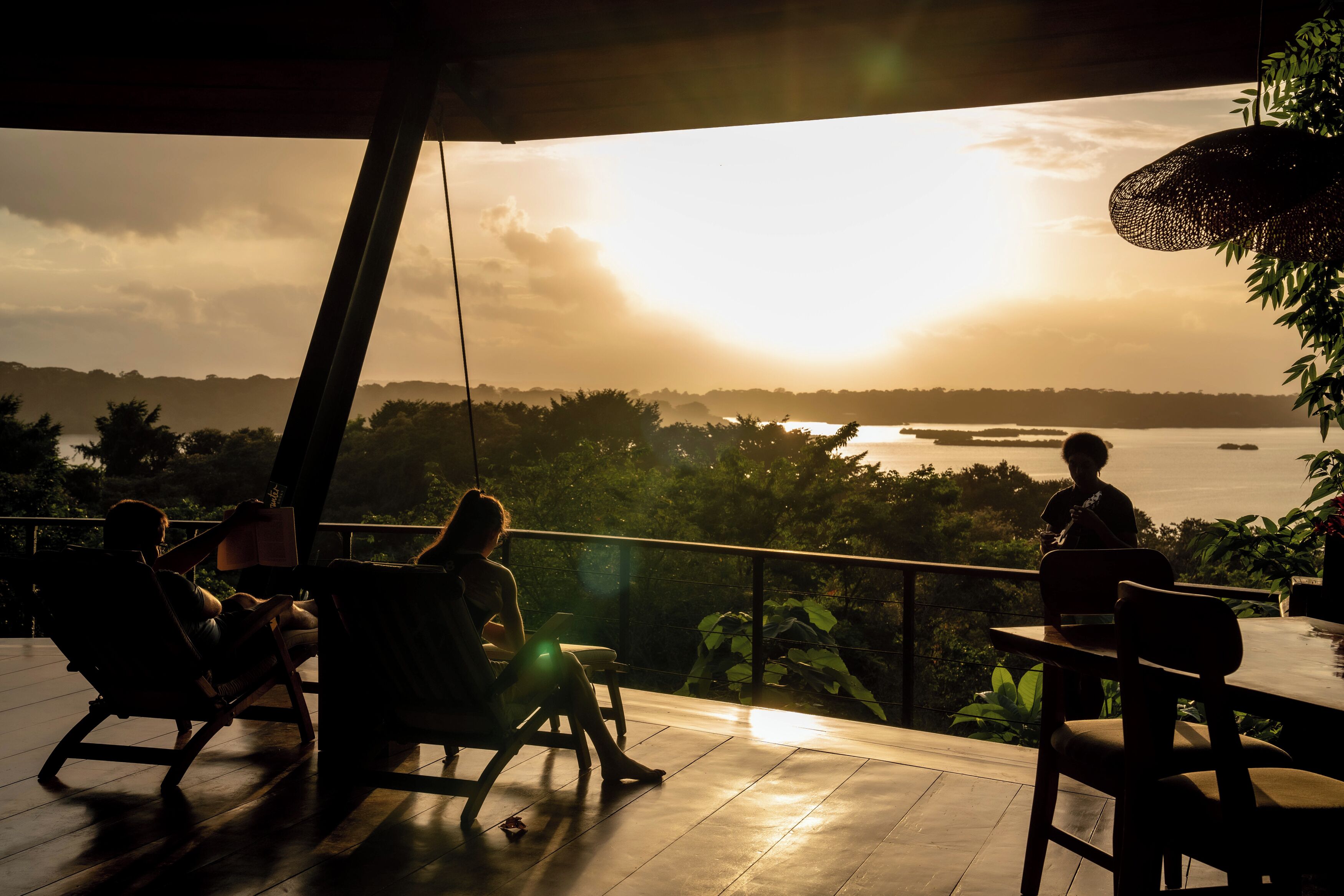 A terrace with a view of the sunset over a body of water, surrounded by greenery.