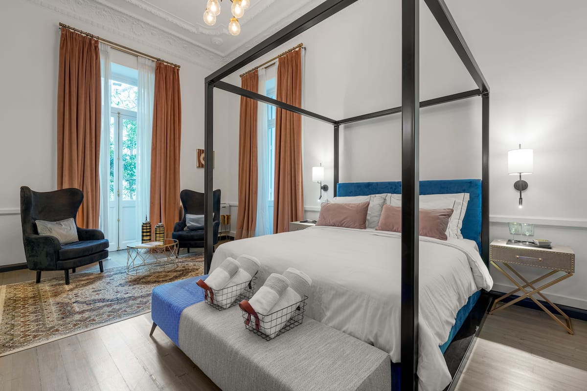 A four-poster bed with white bedding, a blue headboard, and a nightstand with a lamp. There are two armchairs with cushions, a patterned rug, and a bench with towels.