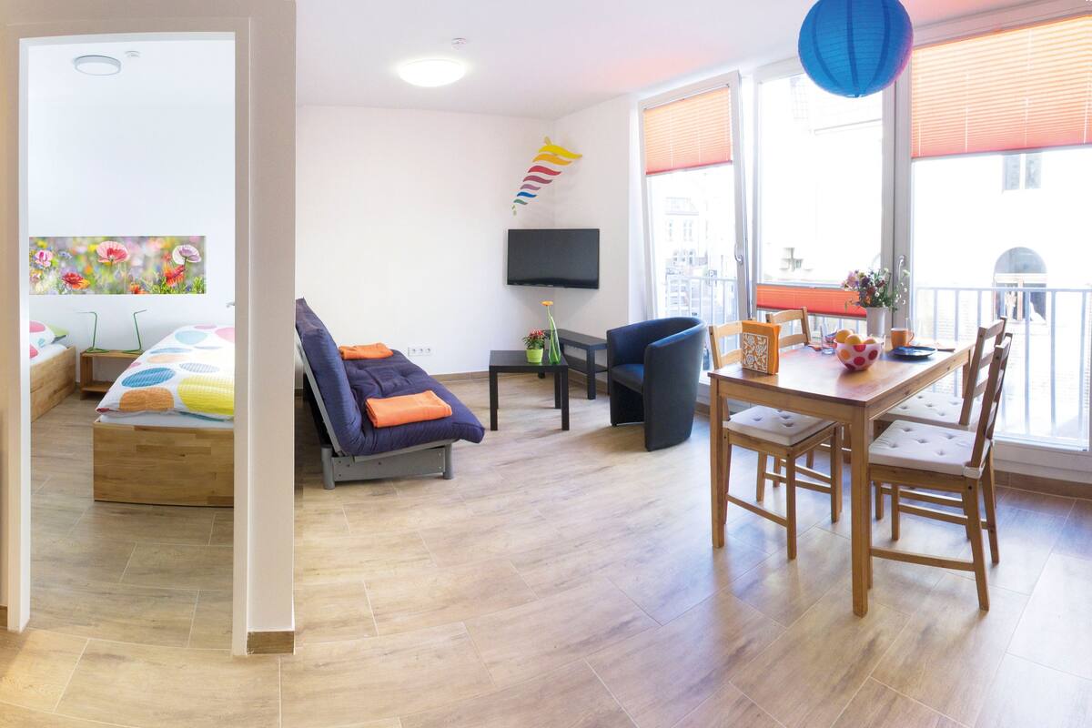 A modern hotel room with a bed, a sofa, a dining table with chairs, a TV, and a balcony with a view.