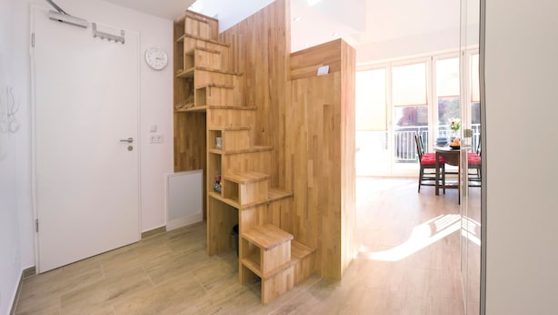 Un interior moderno con una escalera de madera, una puerta blanca y un balcón con mobiliario exterior.