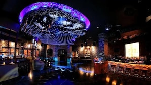 Un espace bar doté d’un grand plafond illuminé, de tabourets de bar et d’une scène où se produit un artiste.