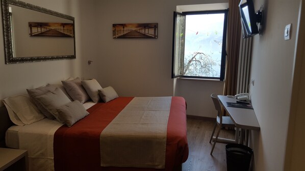 A hotel room with a bed, a desk, a chair, a TV, and a window with a view of trees.
