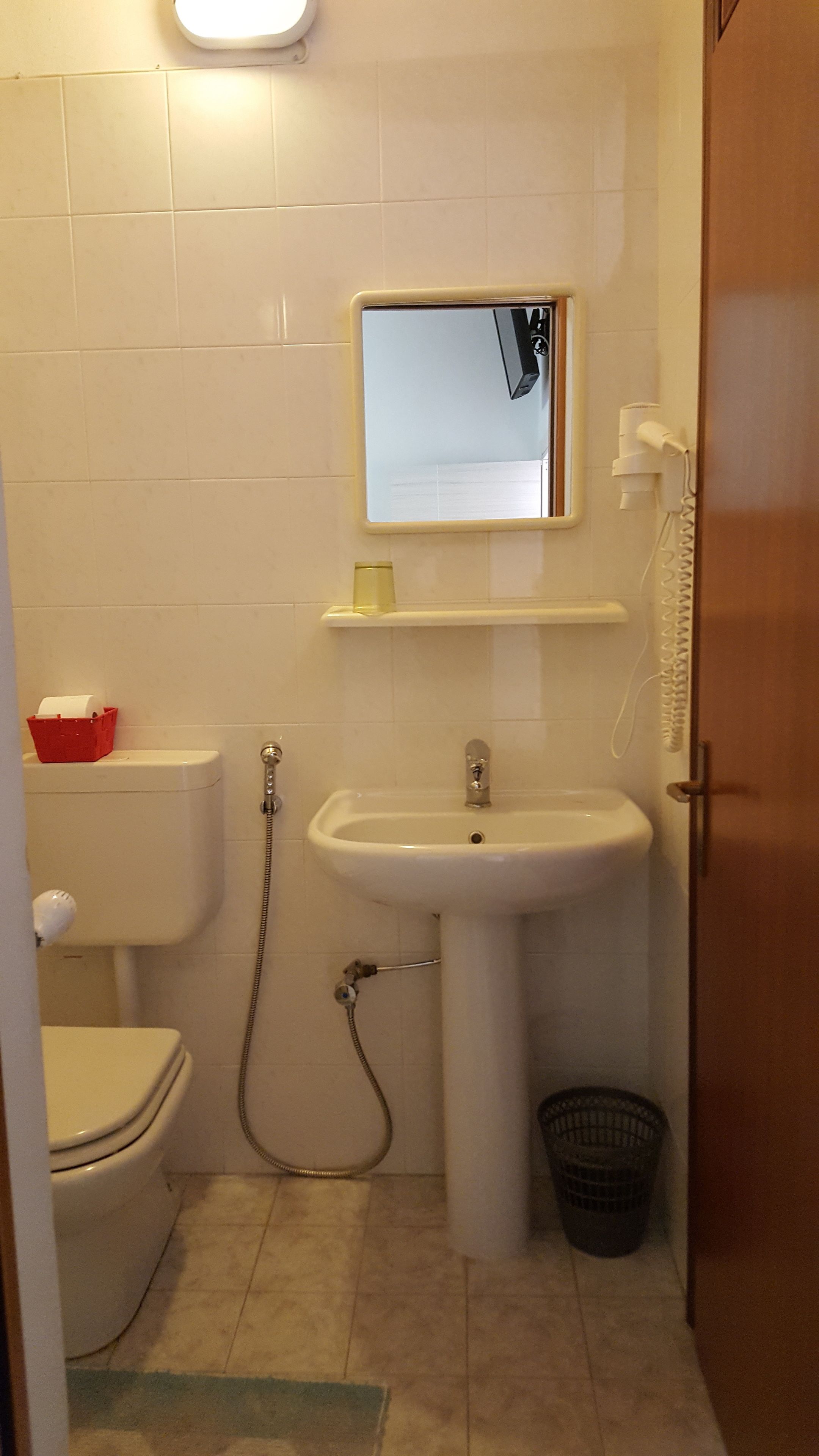 A small bathroom with a toilet, sink, and shower. There is a red basket on the toilet, a white trash bin, and a mirror above the sink.