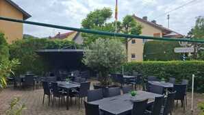 En udendørs terrasse med flere borde og stole, omgivet af grøn vegetation og bygninger.