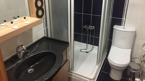 Une salle de bain avec un lavabo noir, une douche en verre, des toilettes blanches et une étagère en bois où sont rangés les articles de toilette.