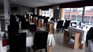 Un restaurant doté de nombreuses tables et chaises, d’un espace cuisine et de grandes fenêtres offrant une vue sur les bâtiments environnants.