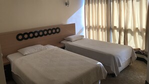 Une chambre d’hôtel avec deux lits simples, une tête de lit en bois, un appareil de climatisation, une lampe fixée au mur et une grande fenêtre avec des rideaux.