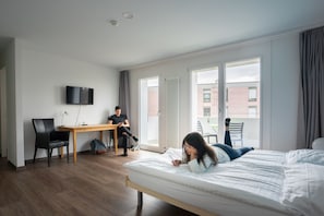 A hotel room with a bed, a desk, a chair, a television, and a view of the outside. - Nyon Hostel (Nyon)