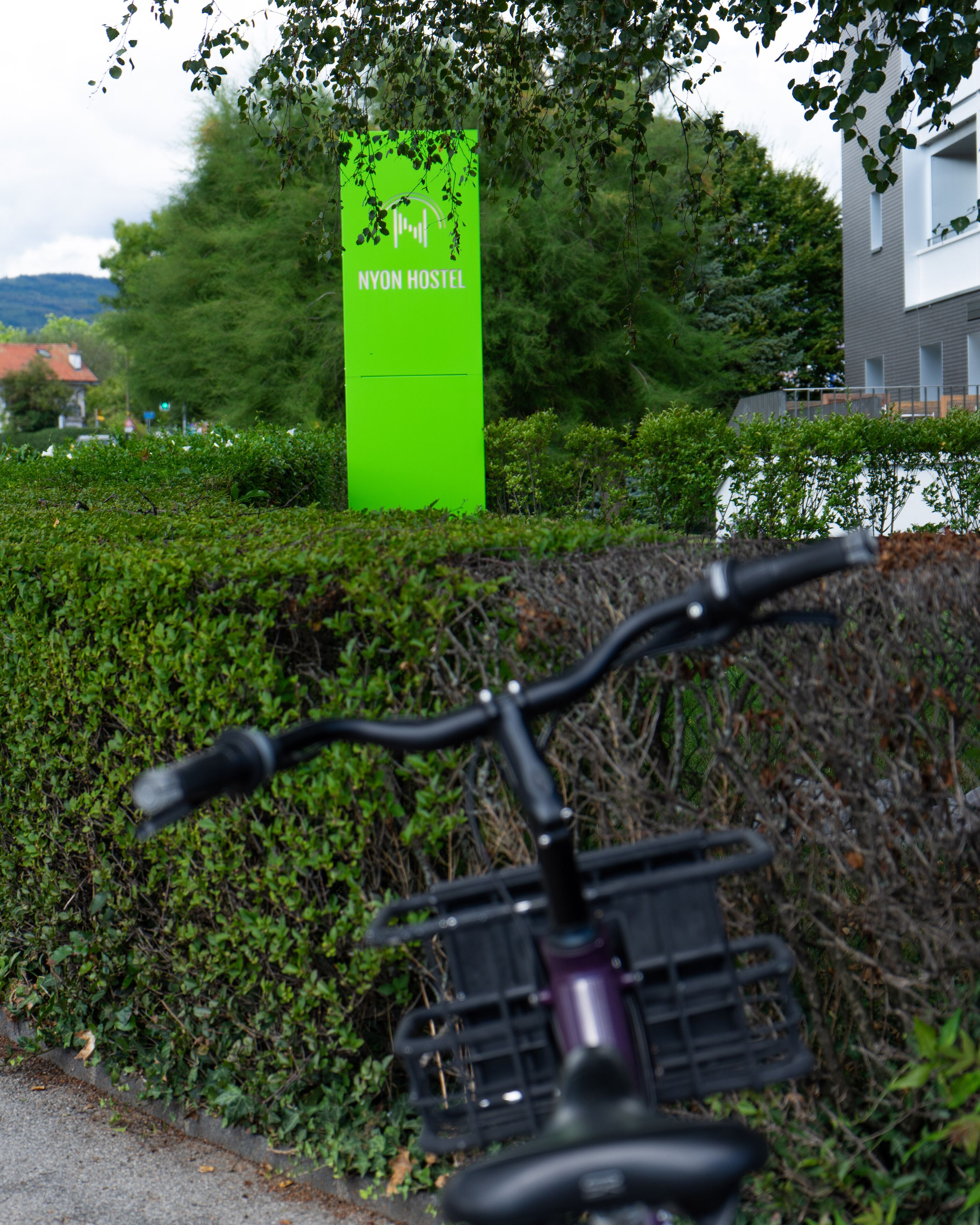 Un vélo est garé devant un panneau vert sur lequel on peut lire : « NYON HOSTEL ».
