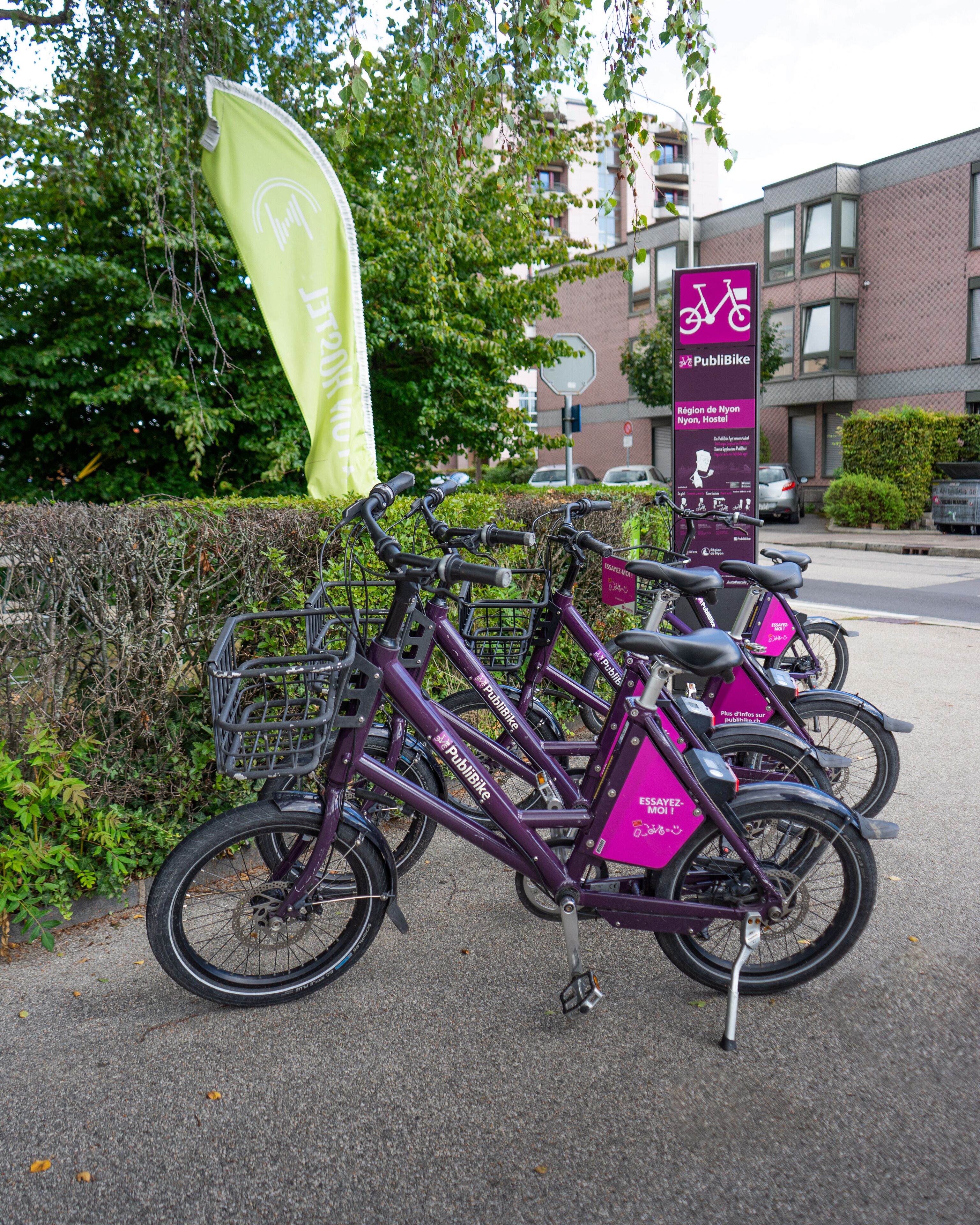 Una fila de bicicletas moradas estacionadas frente a un edificio con un cartel que dice 'Public Bike' y 'Région de Nyon, Nyon Hostel'.