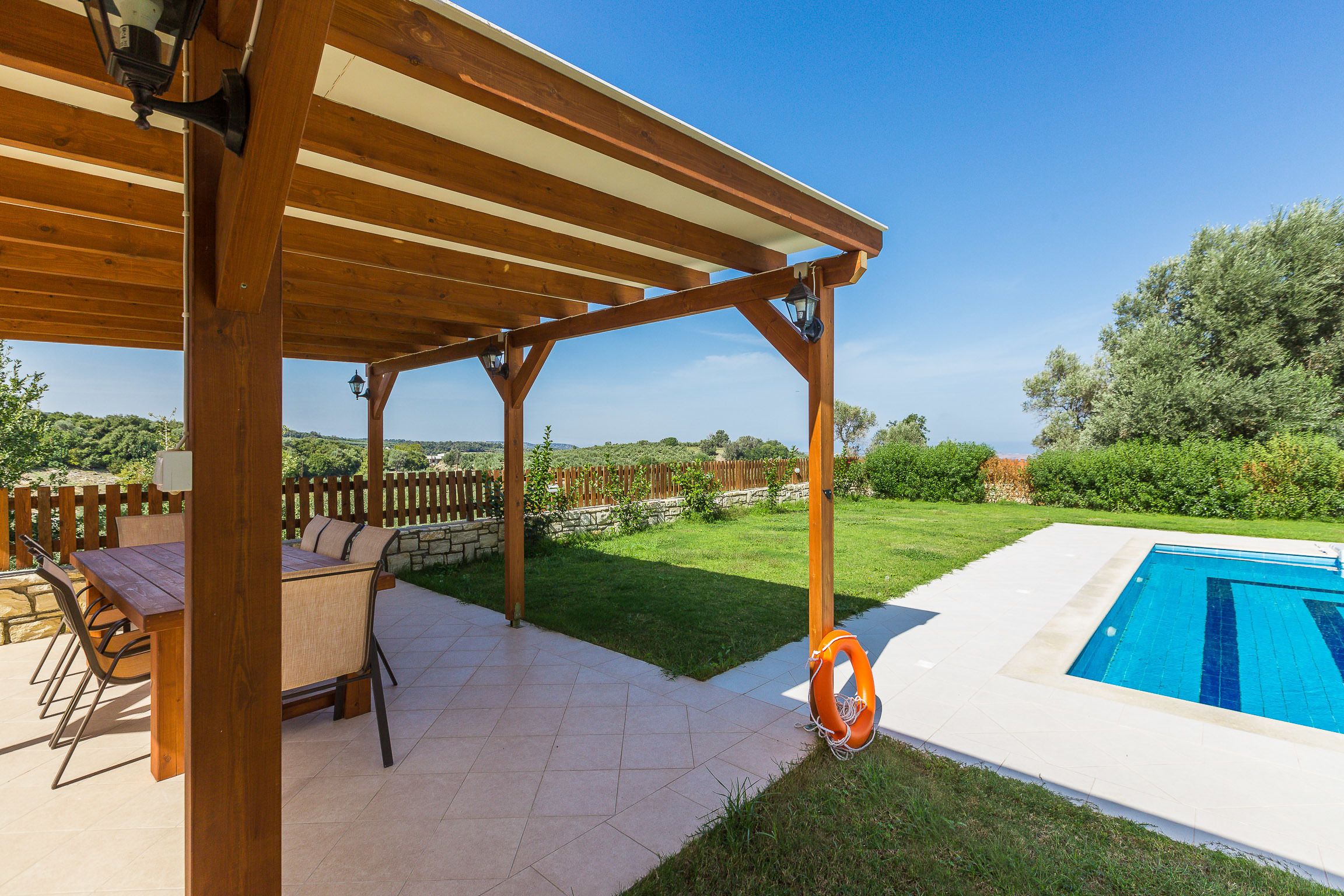 A wooden pergola with a pool table and chairs underneath, a swimming pool, and a grassy yard with a wooden fence.