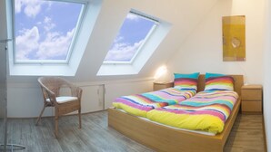 Un dormitorio con tragaluz, una cama con manta a rayas de colores, una silla de mimbre y una mesita de noche con lámpara.