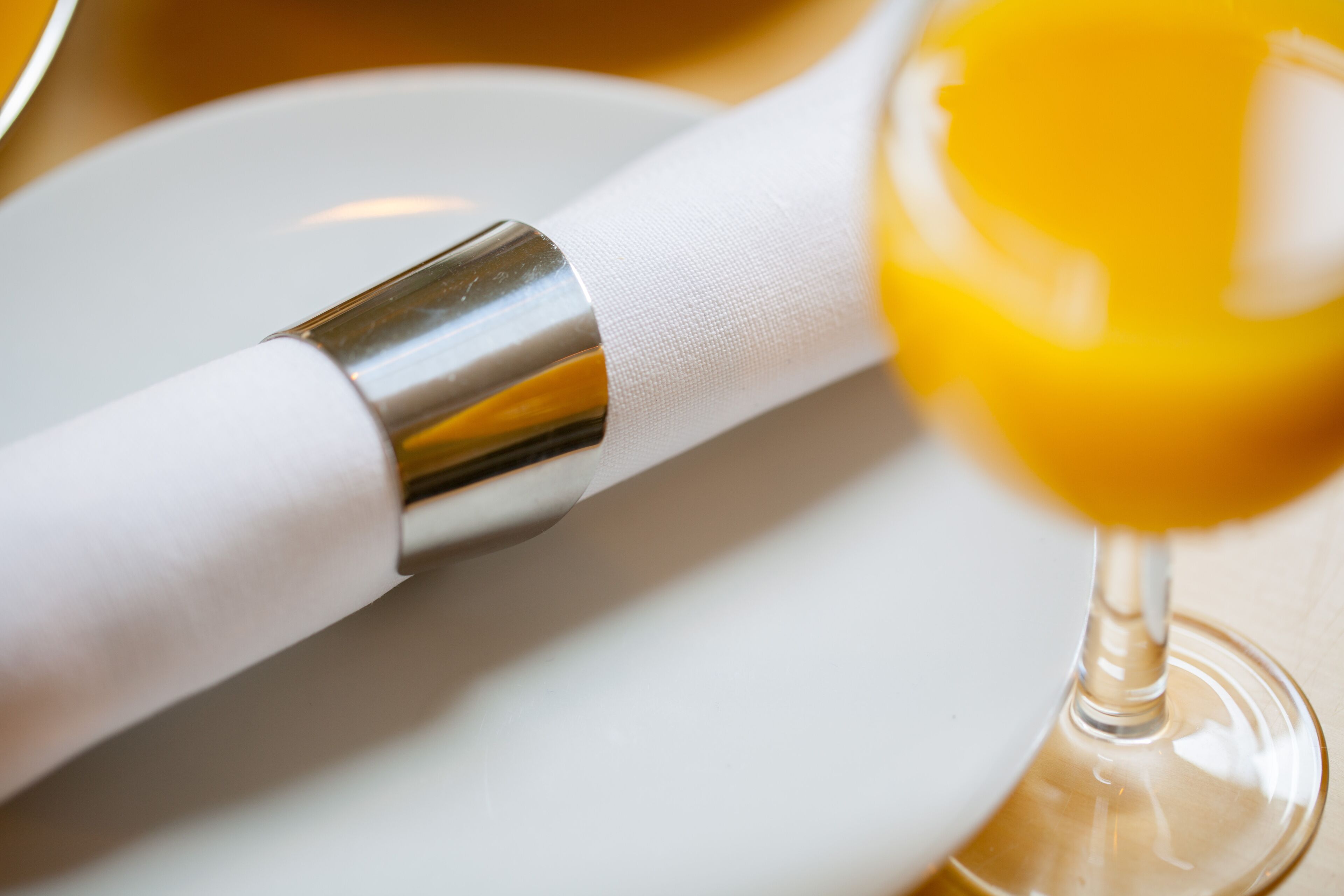 A close-up of a white napkin wrapped around a silver utensil, possibly a spoon or fork, with a glass of yellow liquid, likely a beverage, in the foreground.