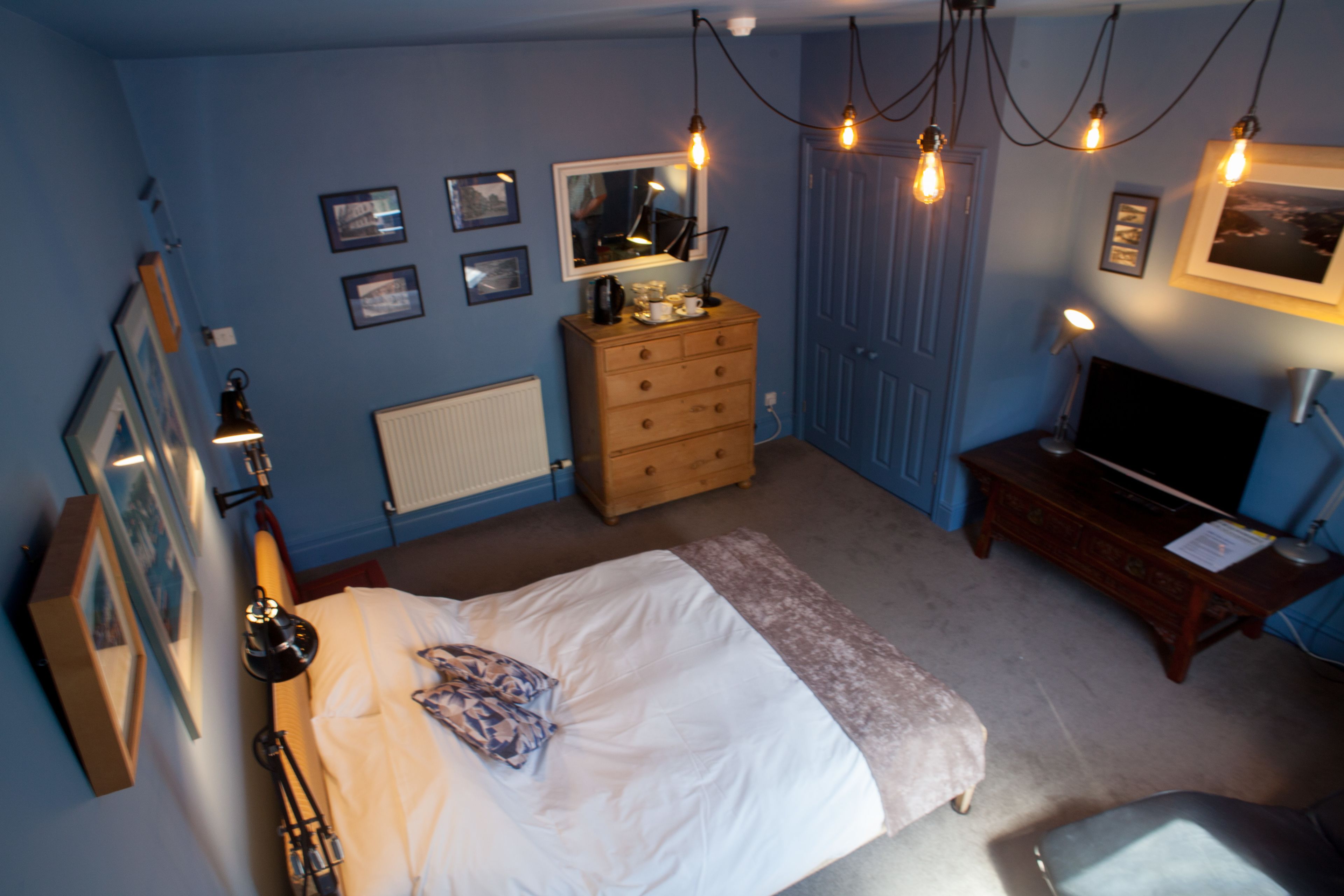A bedroom with a bed, dresser, and TV. There are wall-mounted lights and framed pictures.