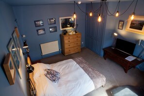 Een slaapkamer met een bed, een dressoir en een televisie. Er zijn wandlampen en beelden in lijsten.