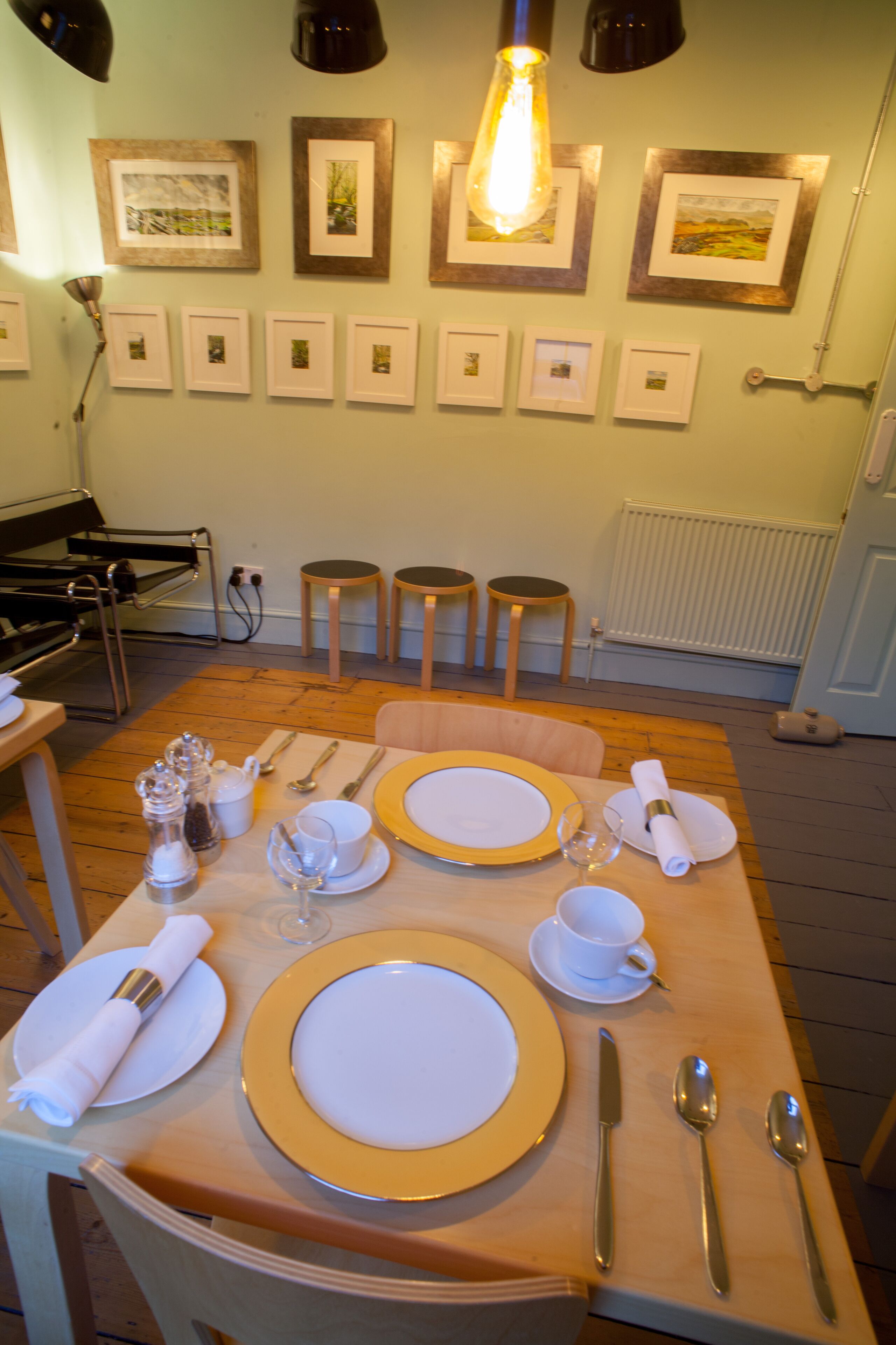 A dining area with a wooden table set for four, including plates, cutlery, and glasses. There are framed pictures on the wall and a radiator below.