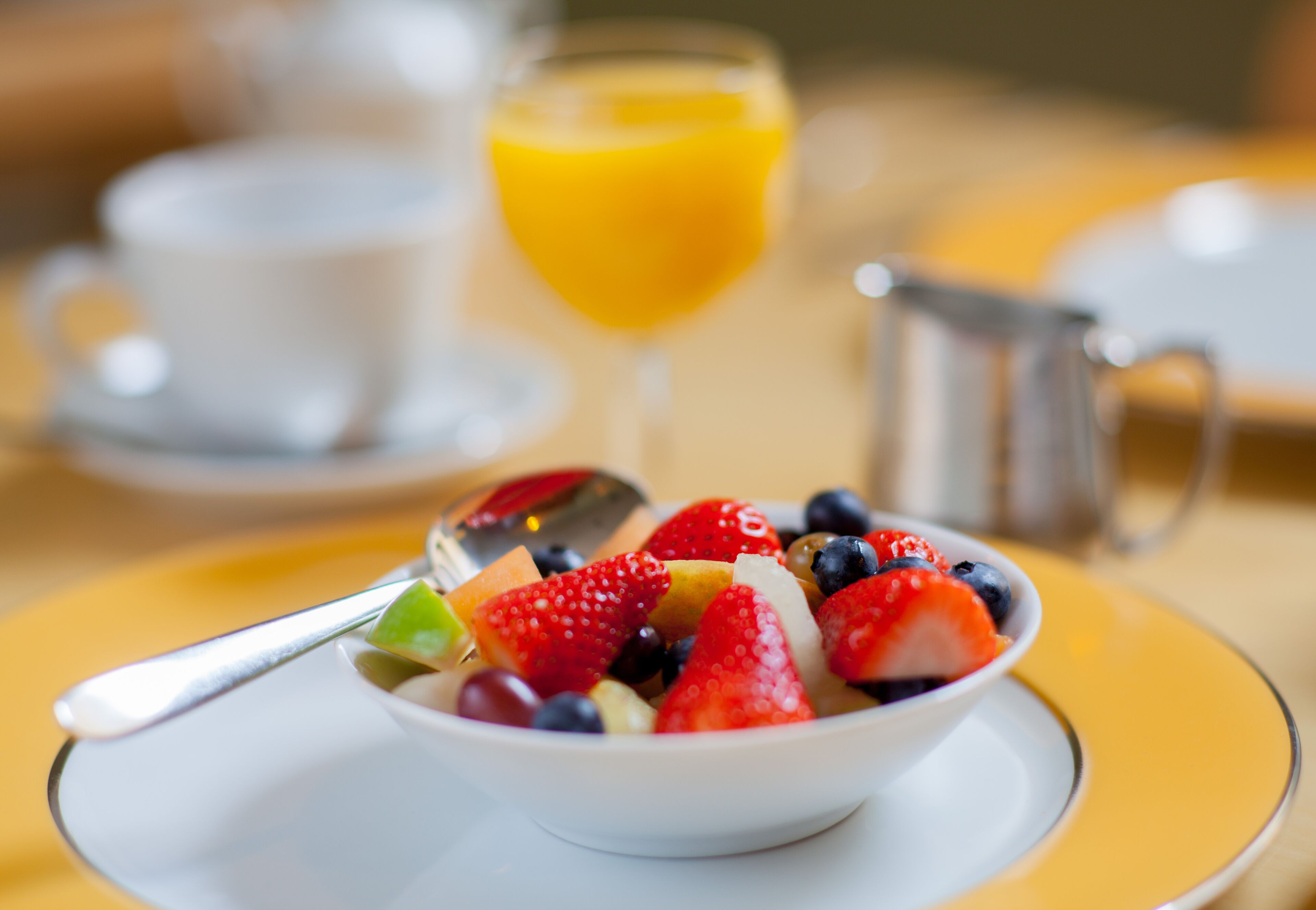 A bowl of fruit salad on a white plate with a spoon, a glass of orange juice, and a white cup in the background.