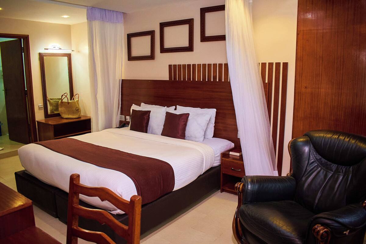 A hotel room with a large bed, a wooden nightstand, a leather armchair, and a mirror on the wall.