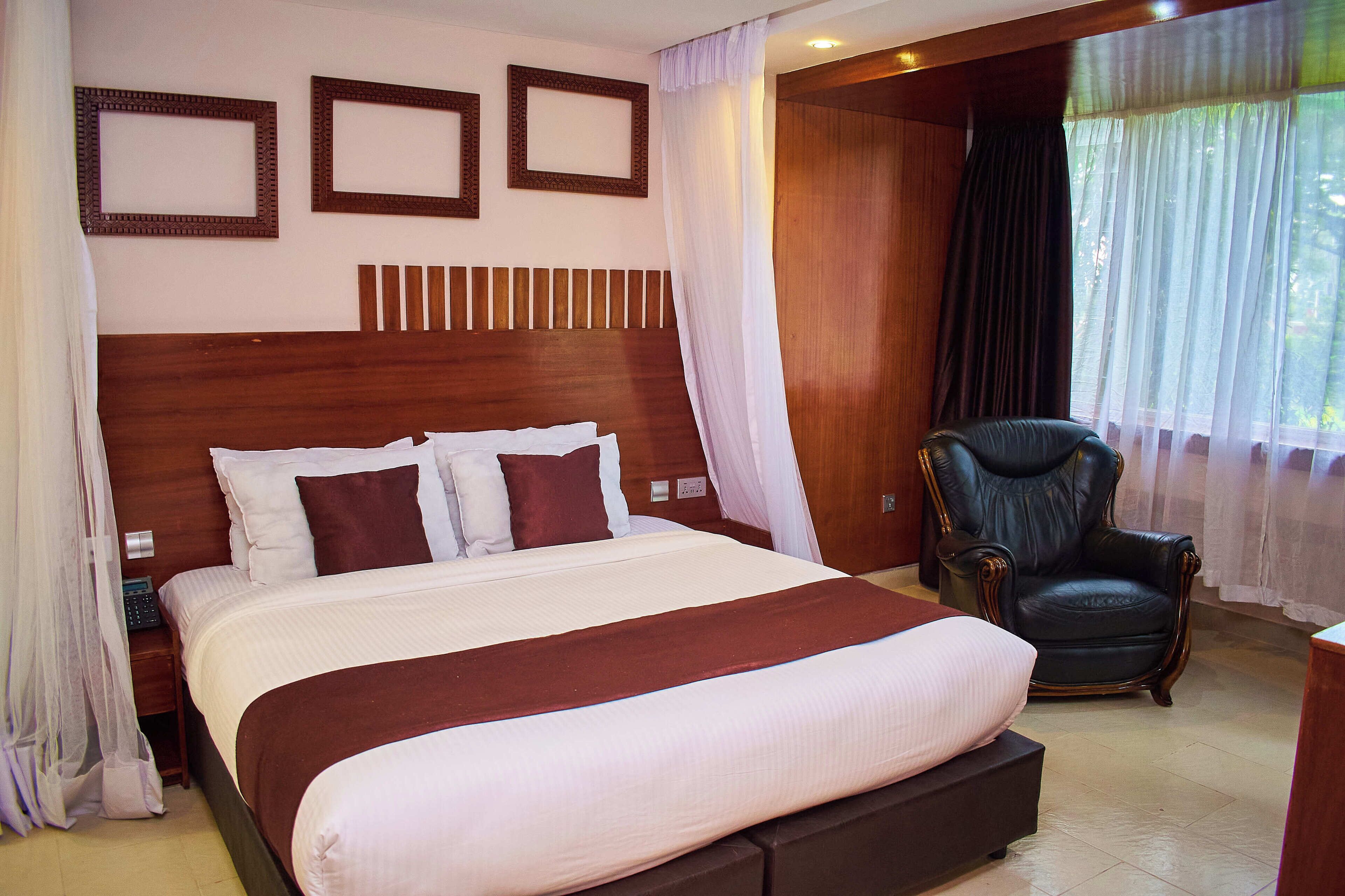 A neatly made bed with a wooden headboard, a black leather chair, and a window with sheer curtains.