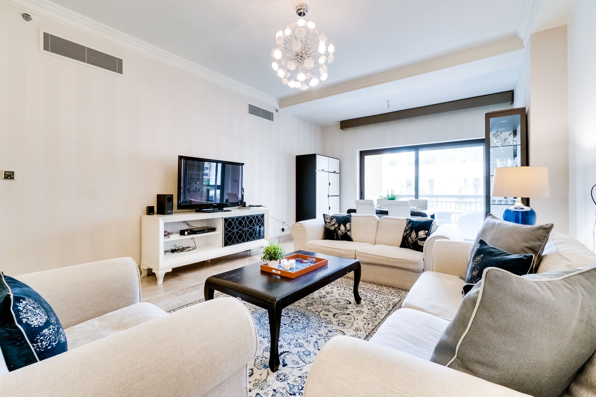 Ein modernes Wohnzimmer mit Flachbildfernseher, weißen Sofas, einem Couchtisch und einem großen Fenster mit Blick ins Freie.