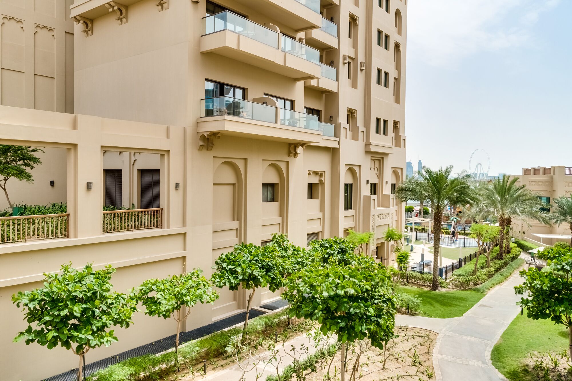 A multi-story residential building with balconies, a landscaped garden, and a clear view of a distant Ferris wheel.