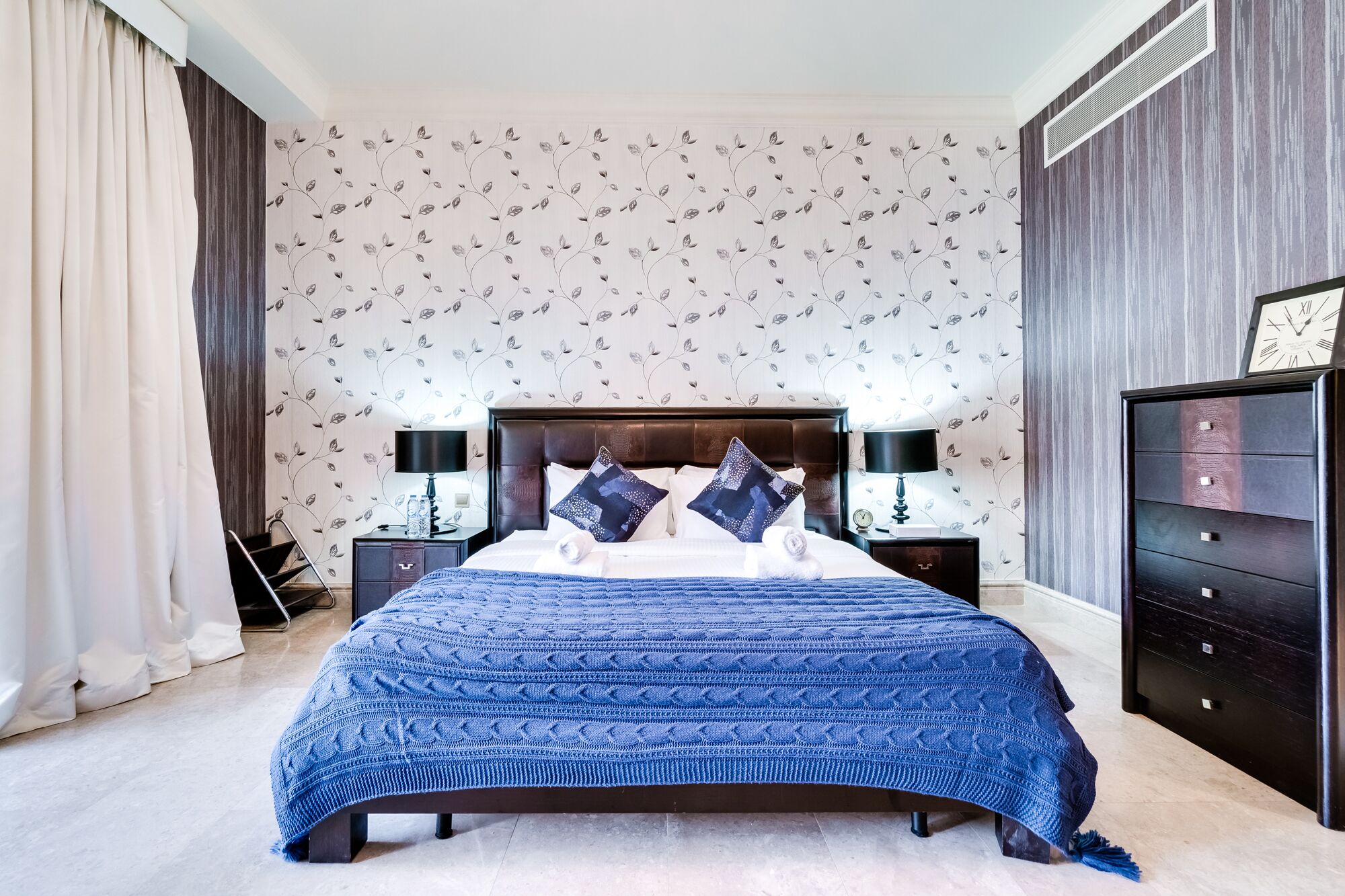A bedroom with a large bed, dark wooden headboard, two bedside tables with lamps, a dresser, and a wall clock.