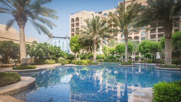 A swimming pool with tiled edges, surrounded by palm trees and greenery, with a multi-story building in the background. - Fairmont North Luxury 2 BR Apt Palm (Dubai)