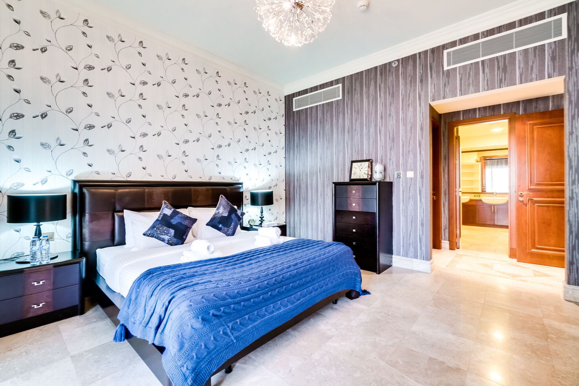 A modern bedroom with a large bed, a nightstand with a lamp, a dresser, and a wardrobe.