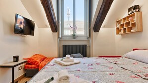 Un dormitorio con televisor, ventana con vistas, cama con ropa de cama floral y estante con utensilios de cocina.
