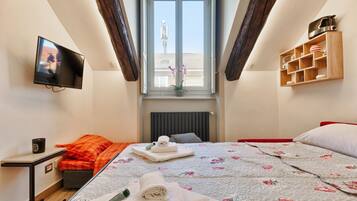 Un dormitorio con televisor, ventana con vistas, cama con ropa de cama floral y estante con utensilios de cocina.