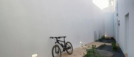 A narrow courtyard with a bicycle leaning against a wall, a small garden area with gravel and plants, and a building with a white facade and a small tower.
