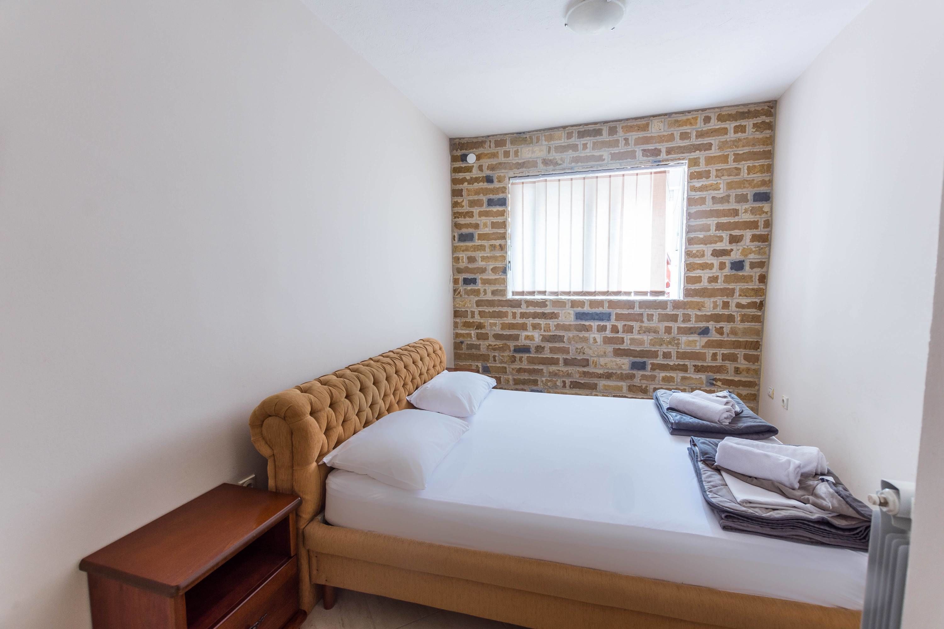 A bedroom with a brick wall, a window with blinds, a bed with white linens, a wooden nightstand, and a folded towel on the bed.