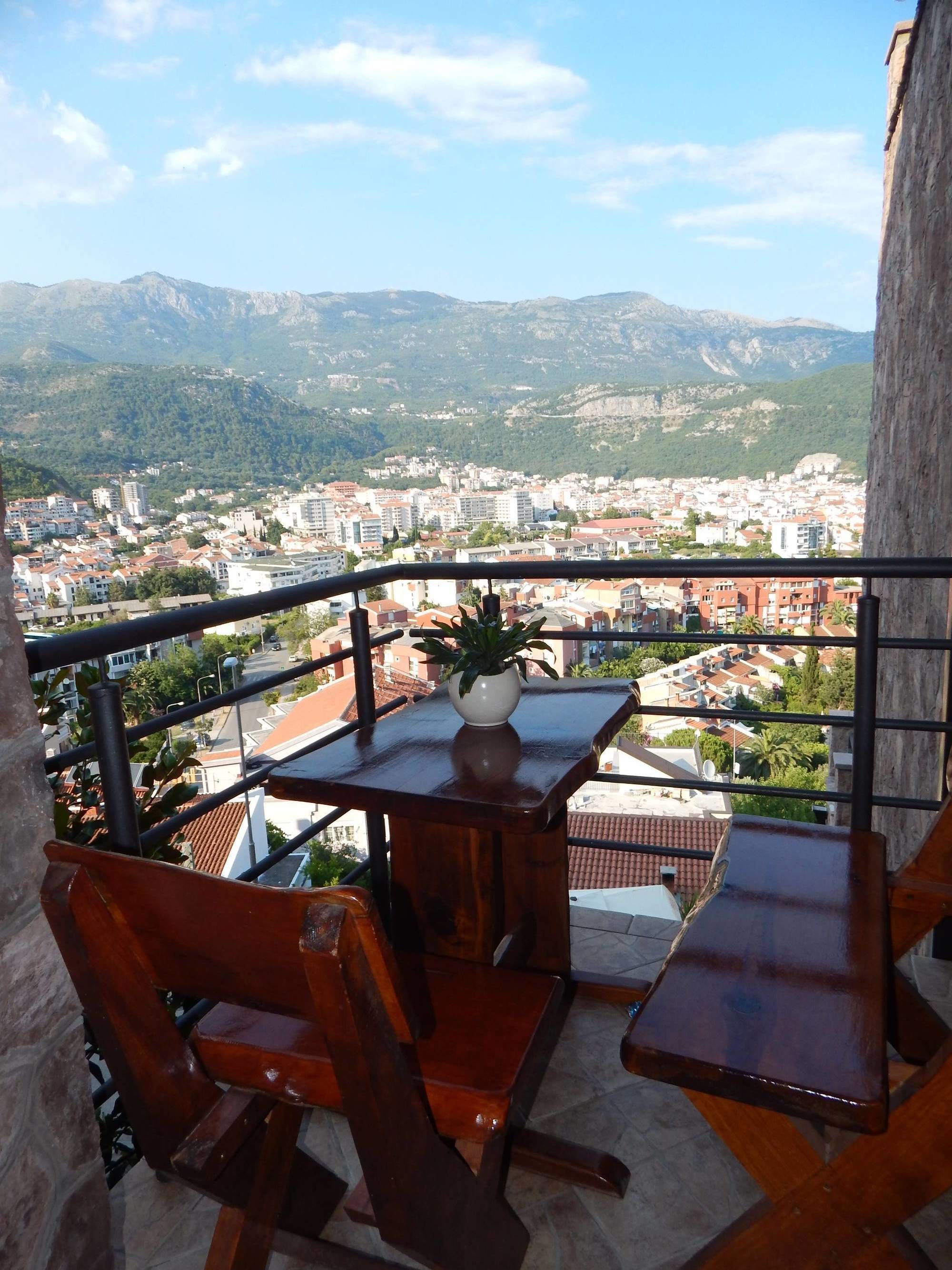 A balcony with wooden furniture, a potted plant, and a view of a cityscape with mountains in the background.