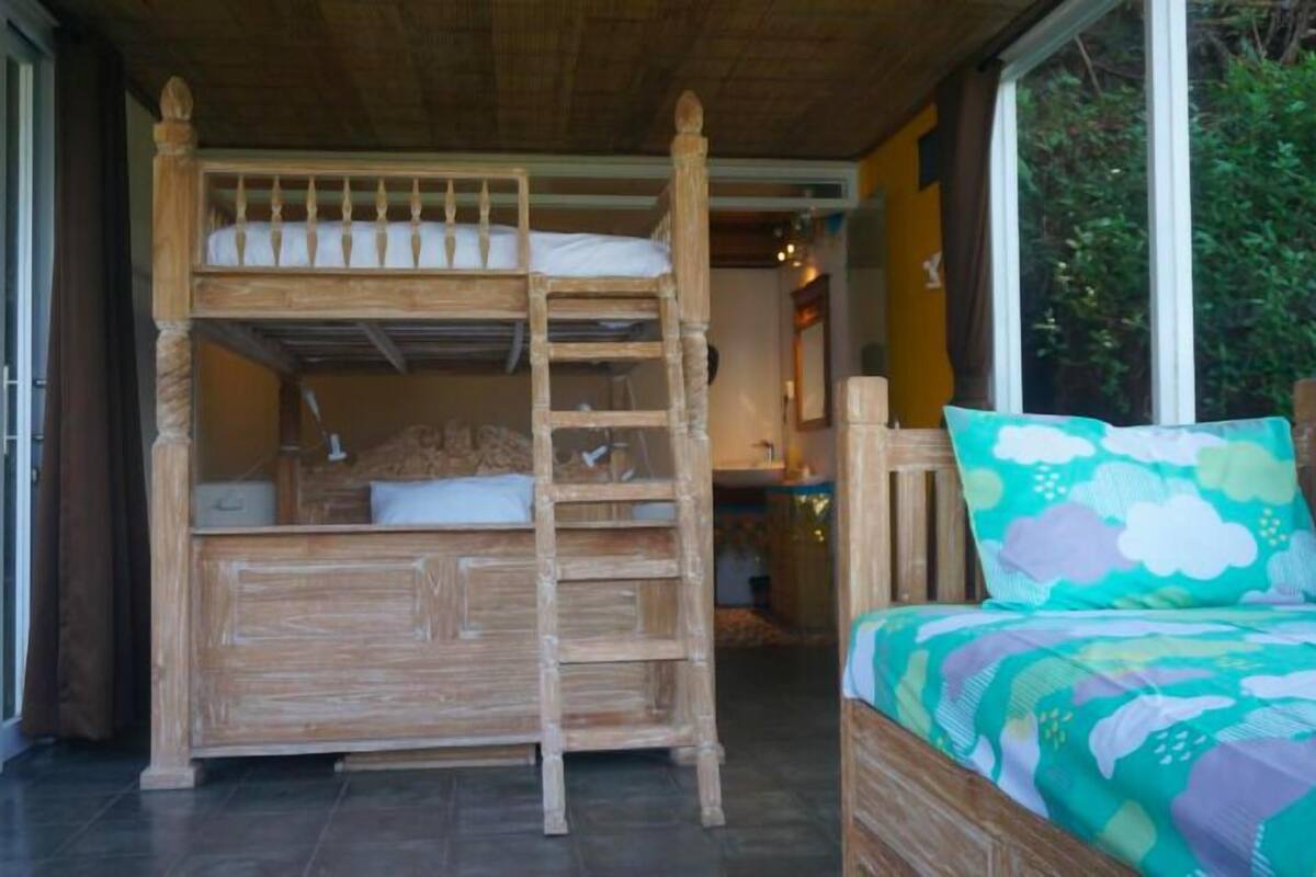 Una cama litera de madera con una escalera, una mesita de noche pequeña y un cojín colorido.
