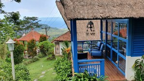 Una estructura de techo de paja con exterior azul, un porche con asientos y vistas del paisaje circundante.