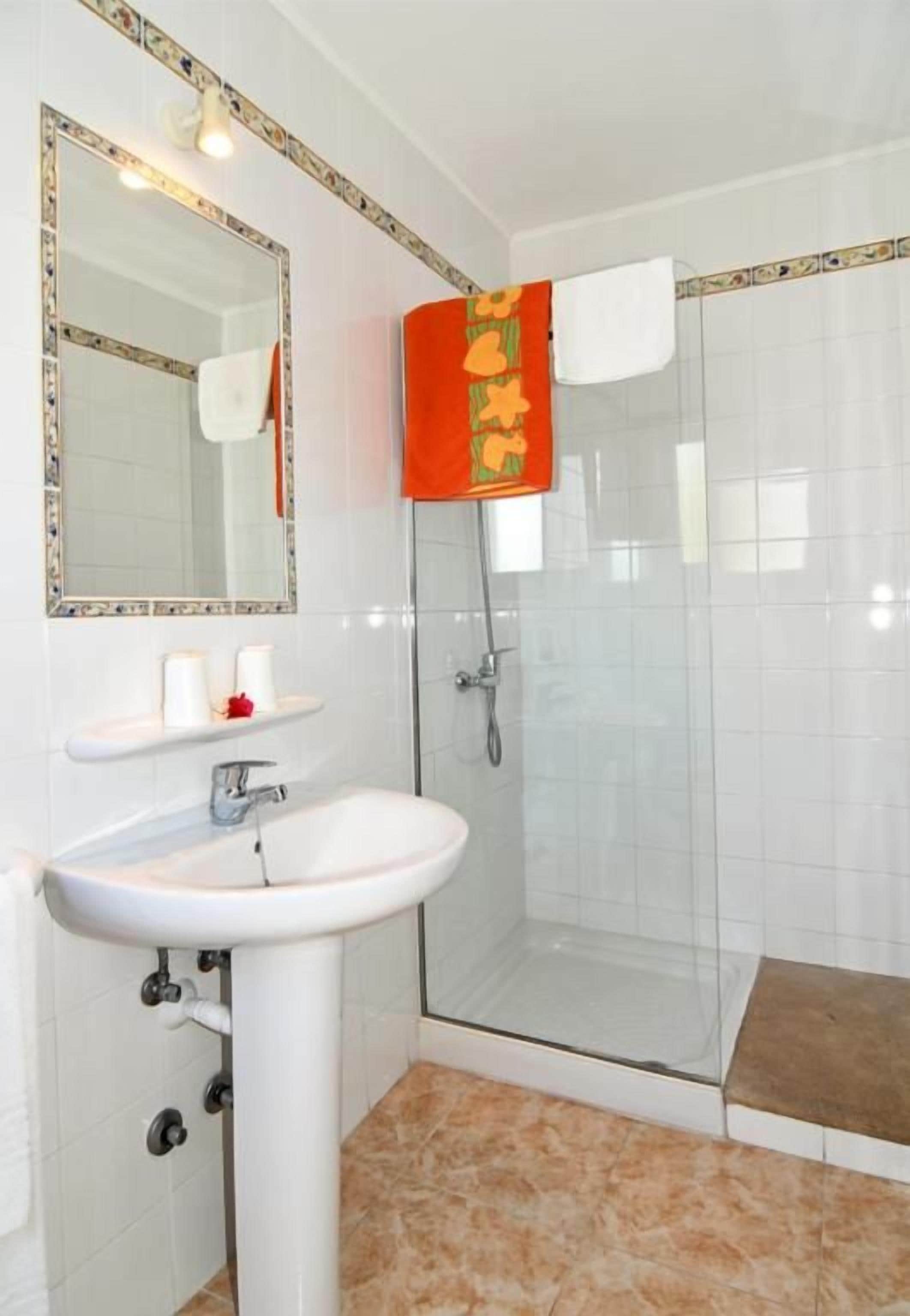 Un baño con un lavamanos blanco, cabina de ducha con mampara de vidrio y espejo con marco decorativo.
