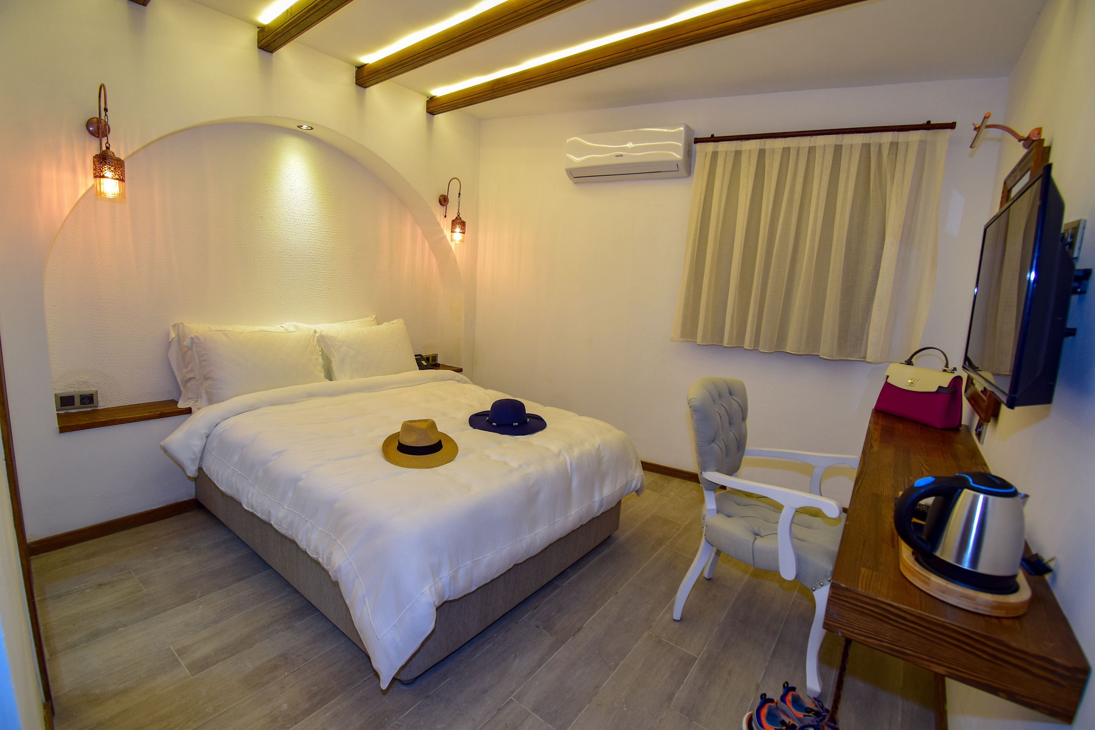 Quarto bem arrumado com cama, abajures, uma cadeira, escrivaninha com chaleira e ar condicionado.