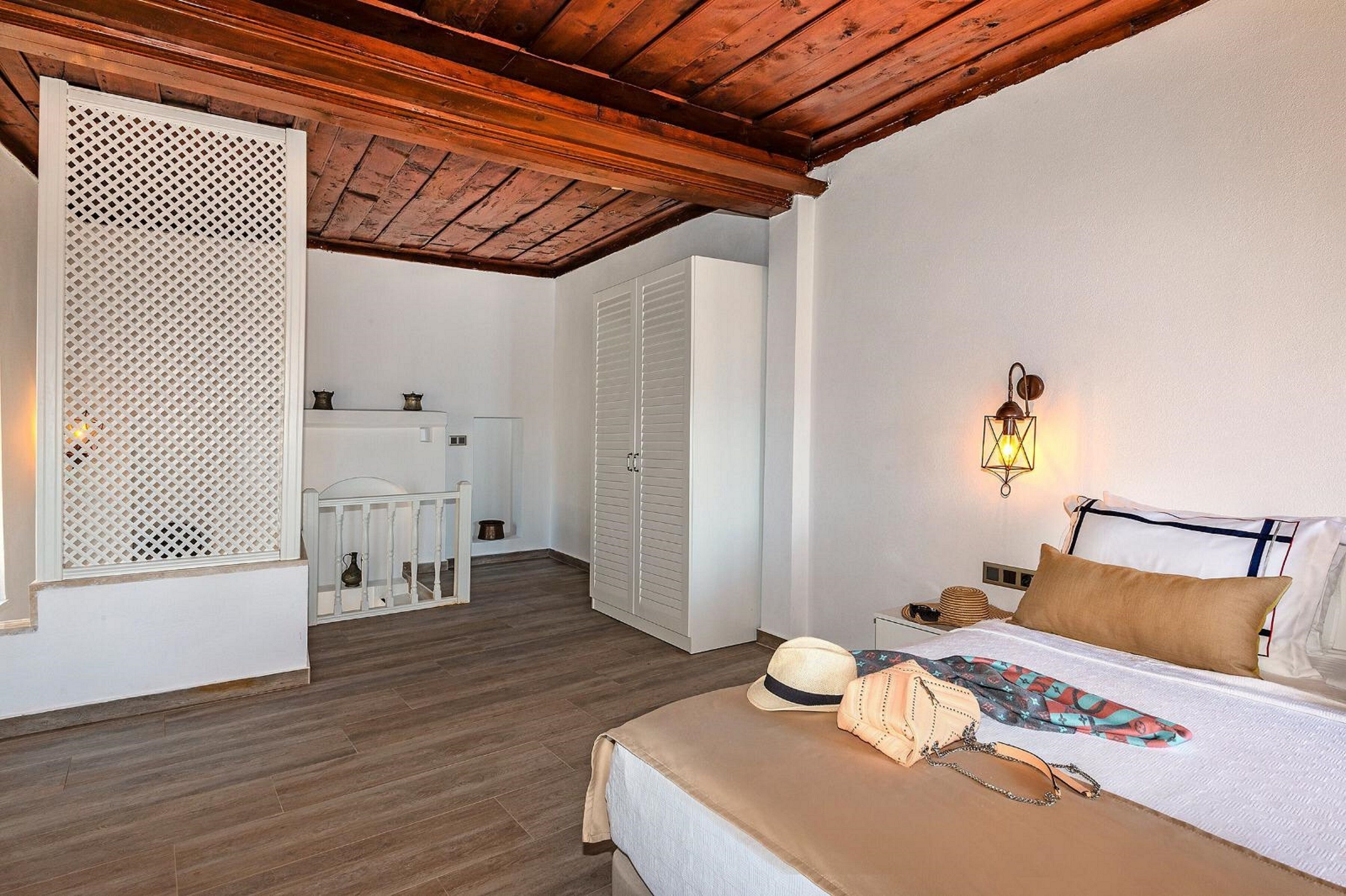 Quarto com teto de madeira, cama com colcha bege, mesa de cabeceira, luminária na parede e guarda-roupa.