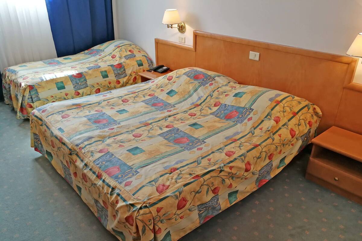 A hotel room with two beds, each with floral-patterned bedding, a wooden headboard, bedside tables, and a wall-mounted lamp.